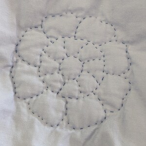 May include: Close-up of a white fabric with a stitched floral design. The design is made of connected, rounded shapes outlined with dark blue thread. The stitching creates a textured, quilted effect.