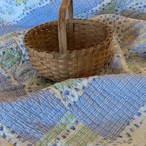 May include: A woven brown basket with a wooden handle. The basket is round and sits on a blue and white floral patterned quilt. The quilt has a patchwork design with various shades of blue, green, and white.