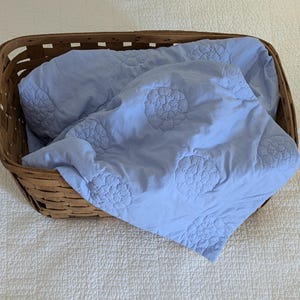 May include: A light blue quilt with floral embroidery rests inside a brown woven basket. The quilt has a soft, textured appearance and is folded neatly. The basket is rectangular and has a rustic, natural look.