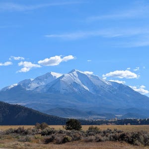 May include: A scenic landscape featuring a snow-capped mountain under a bright blue sky with scattered clouds. The foreground shows a field of dry grass and shrubs, with a dark green forested area on the left.