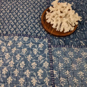 May include: A quilted textile featuring a patchwork of blue and white floral and paisley patterns, with a white branching coral specimen resting on a circular dark brown wooden base placed directly on the surface of the fabric.