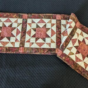 May include: A rectangular quilted table runner featuring a repeating star pattern in shades of dusty rose, cream, and mottled brown. The fabric displays a textured, marbled appearance with geometric pieced blocks arranged in a linear sequence.