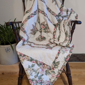 May include: A white quilt with a floral pattern and star-shaped designs draped over a wooden chair. The quilt features pink and green roses and leaves along the edges and in the star patterns. The chair is dark brown.