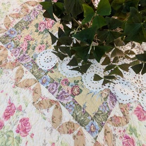 May include: A quilted fabric featuring a floral pattern with pink roses, blue flowers, and green leaves on a cream background, accented by tan leaf-shaped appliques and white crocheted doilies, with a dark green shamrock plant on top.