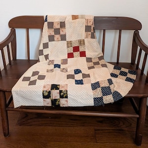 May include: A vintage quilt with a patchwork design in shades of cream, brown, red, and blue, draped over a wooden bench. The quilt features a repeating pattern of squares and floral prints. The bench has a curved back and armrests.