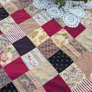 May include: A patchwork quilt with a variety of patterned fabrics in red, cream, black, and tan. The quilt features floral, striped, and paisley designs. A white lace doily and a wicker basket with greenery are in the background.
