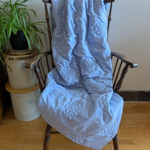 May include: A light blue quilt with embroidered floral patterns draped over a wooden chair. The quilt's design features repeating circular floral motifs. The chair has a dark wood finish and a curved back. A potted plant is visible in the background.