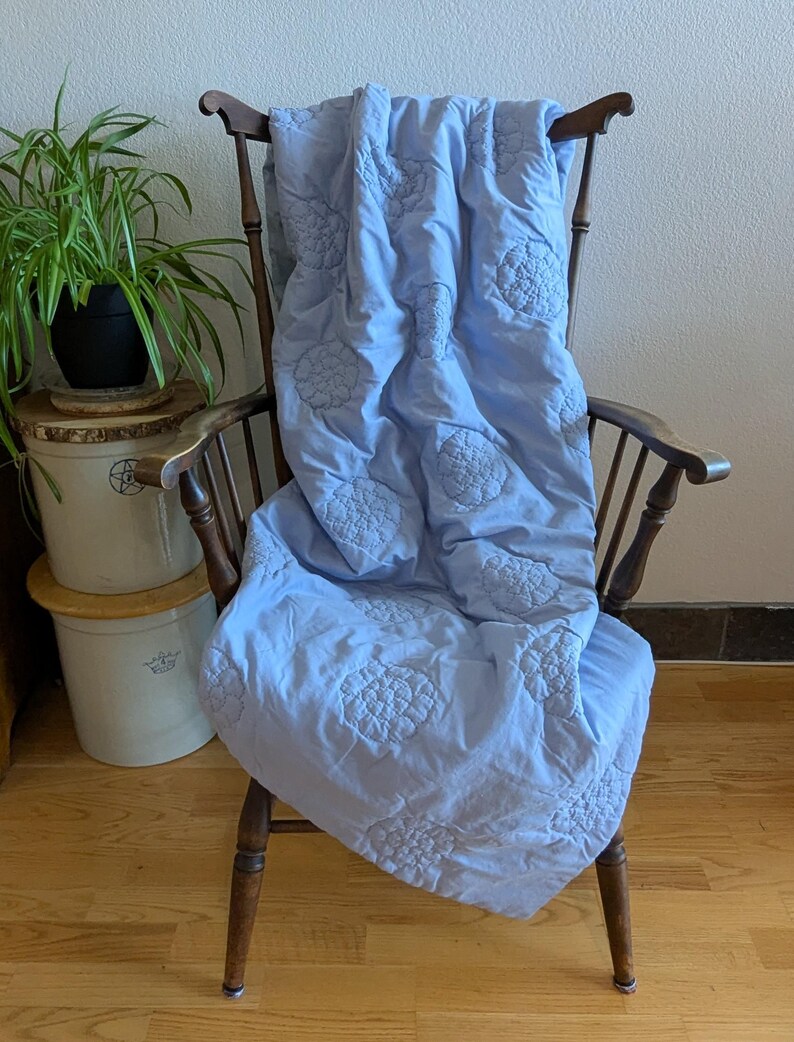 May include: A light blue quilt with embroidered floral patterns draped over a wooden chair. The quilt's design features repeating circular floral motifs. The chair has a dark wood finish and a curved back. A potted plant is visible in the background.