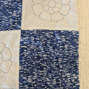 May include: Close-up of a quilt with a floral design. The quilt features alternating squares of white fabric with embroidered flowers and blue patterned fabric. The stitching is in dark gray thread, and the overall aesthetic is a mix of traditional and modern.