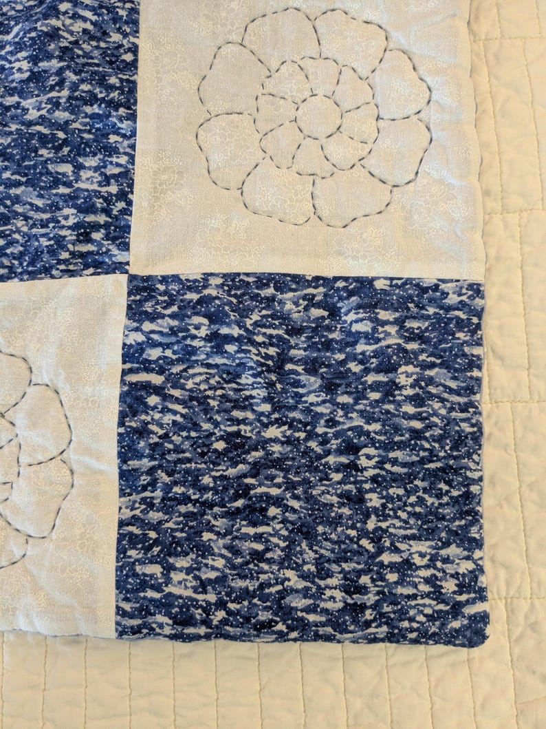 May include: Close-up of a quilt with a floral design. The quilt features alternating squares of white fabric with embroidered flowers and blue patterned fabric. The stitching is in dark gray thread, and the overall aesthetic is a mix of traditional and modern.