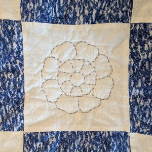 May include: Close-up of a quilted textile with a floral design. The quilt features a white center with a stitched flower and is framed by blue and white patterned squares. The stitching is in a dark gray color.