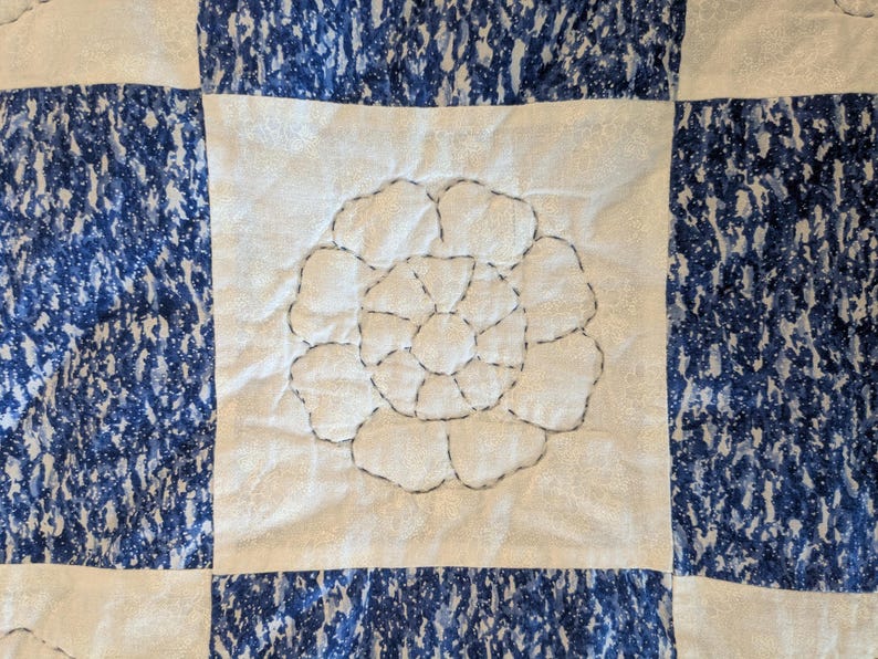 May include: Close-up of a quilted textile with a floral design. The quilt features a white center with a stitched flower and is framed by blue and white patterned squares. The stitching is in a dark gray color.