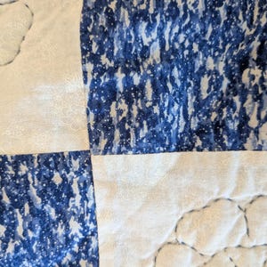 May include: Close-up of a quilted fabric with a patchwork design. The quilt features alternating squares of white fabric with a floral pattern and blue fabric with a speckled pattern. The stitching is visible.