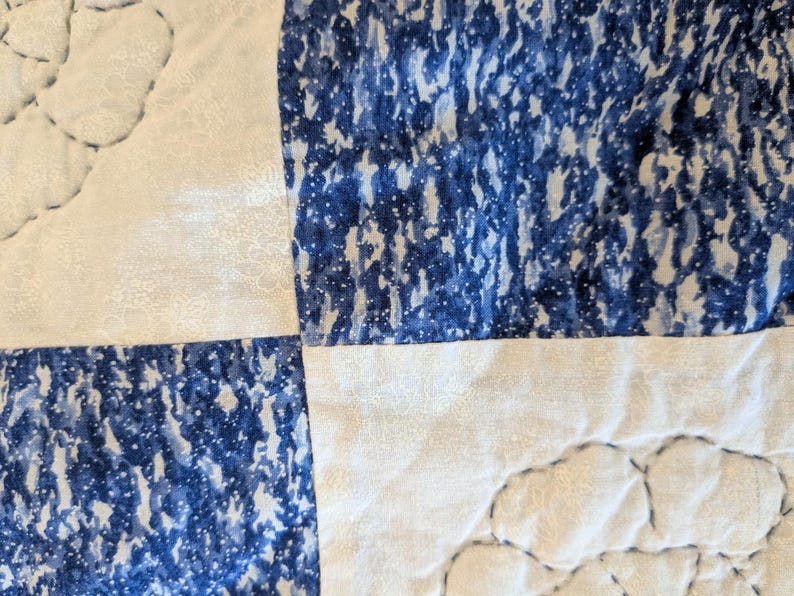 May include: Close-up of a quilted fabric with a patchwork design. The quilt features alternating squares of white fabric with a floral pattern and blue fabric with a speckled pattern. The stitching is visible.