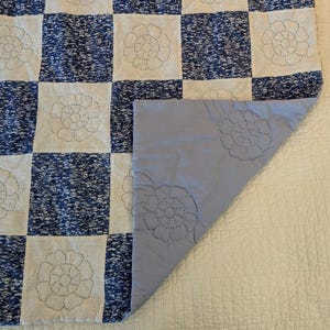 May include: A blue and white patchwork quilt with a floral design. The quilt features alternating squares of blue speckled fabric and white fabric with embroidered floral patterns. The underside is a solid light blue color with matching floral embroidery.
