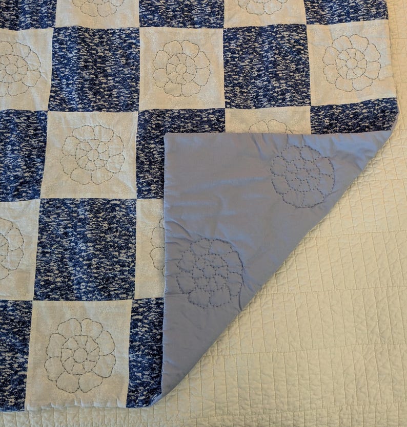 May include: A blue and white patchwork quilt with a floral design. The quilt features alternating squares of blue speckled fabric and white fabric with embroidered floral patterns. The underside is a solid light blue color with matching floral embroidery.
