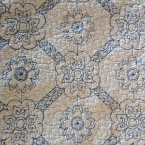 May include: Close-up of a quilted fabric with a repeating floral pattern. The design features embroidered blue flowers and geometric lines on a cream-colored background. The texture appears soft and slightly raised.