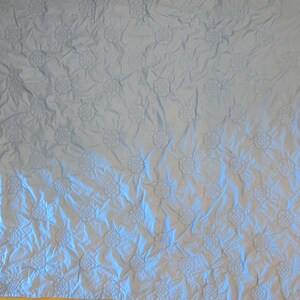 May include: A light blue quilt with a repeating floral pattern. The flowers are raised, creating a textured surface. The quilt has a soft, wrinkled appearance, suggesting it is made of a lightweight fabric. The overall design is simple and elegant.