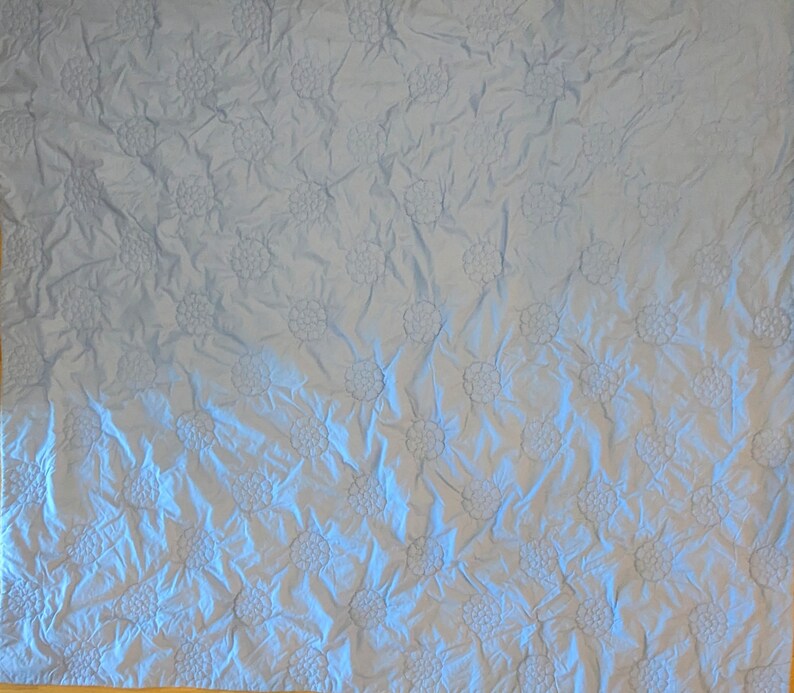 May include: A light blue quilt with a repeating floral pattern. The flowers are raised, creating a textured surface. The quilt has a soft, wrinkled appearance, suggesting it is made of a lightweight fabric. The overall design is simple and elegant.