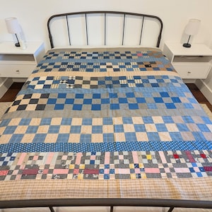 May include: A patchwork quilt with a variety of blue, beige, and gray fabrics. The quilt features checkerboard patterns, polka dots, and other geometric designs. The quilt is on a dark metal bed frame, with white nightstands and lamps on either side.