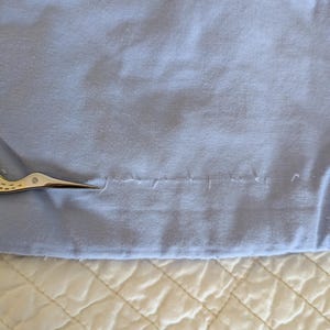 May include: A close-up of a light blue fabric with a seam and small scissors. The fabric is slightly wrinkled and appears to be cotton or a similar material. The scissors are silver and gold.