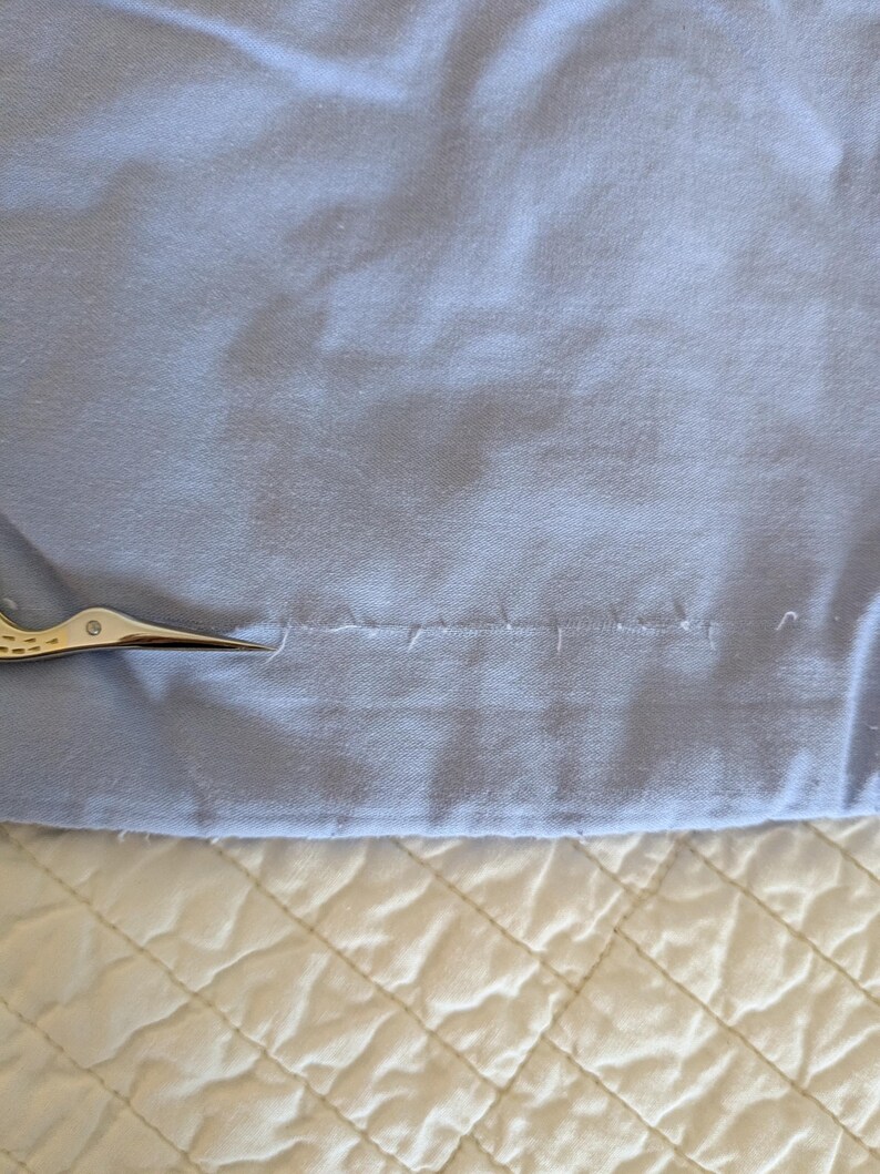May include: A close-up of a light blue fabric with a seam and small scissors. The fabric is slightly wrinkled and appears to be cotton or a similar material. The scissors are silver and gold.