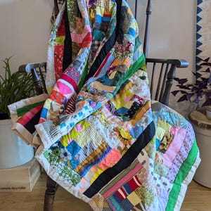 May include: A handmade patchwork quilt draped over a wooden chair. The quilt features a variety of colorful fabric scraps in a mix of patterns and textures, including floral, striped, and solid designs. Black and green borders frame the quilt.