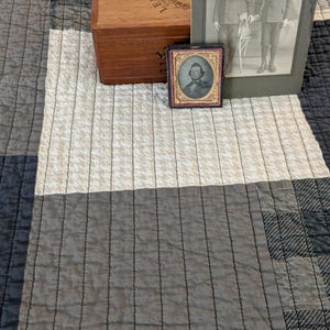 May include: A patchwork quilt featuring alternating squares of cream houndstooth and solid charcoal gray fabric. Resting on the quilt is a wooden Lempira cigar box, a large rectangular black-and-white photograph of two boys, and a small framed portrait.