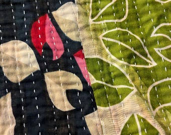 May include: Close-up of a quilted textile with a floral design. The textile features a dark blue section with red and beige leaf shapes, and a green section with white leaf outlines. White stitching is visible.