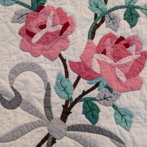 May include: A close-up of a white quilt with a floral design. The quilt features pink roses with red and white accents, brown stems, and green and blue leaves. A gray ribbon is tied in a bow at the base of the flowers.