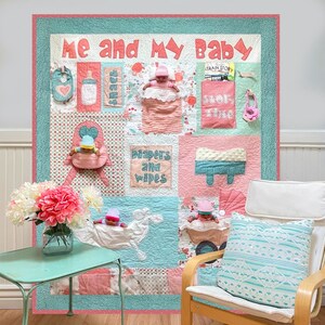 Peut inclure: Une courtepointe en patchwork rose et bleu avec les mots "Moi et mon bébé" en blanc. La courtepointe a différentes poches avec différents articles pour bébés, dont une bouteille, une couche et une sucette. La courtepointe est affichée sur un mur avec une chaise blanche devant.