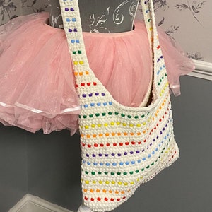 May include: A white crochet bag with a rainbow stripe pattern. The bag has a long strap and is made with colorful beads.