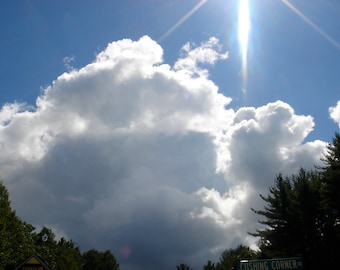 May include: A bright blue sky with fluffy white clouds and a sun shining through the clouds. The sun is shining so brightly that it is creating a starburst effect.