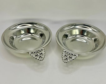 May include: Two silver serving dishes with ornate handles. The dishes are round and have a slightly raised rim.