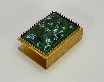 Puede incluir: Una pequeña caja de cerillas de color dorado con una tapa de mármol verde y blanco. La tapa está rodeada por un borde dorado decorativo.