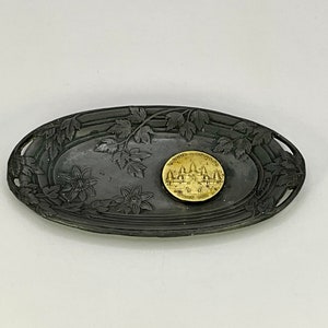 Può includere: Un vassoio ovale in peltro con un design floreale. Il vassoio ha un bordo rialzato e due maniglie ritagliate. Una piccola moneta rotonda dorata con le parole "Women of Courage" e un design di edificio si trova al centro del vassoio.