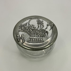 Pode incluir: Um frasco de vidro transparente com uma tampa metálica decorativa. A tampa apresenta um relevo detalhado de uma cesta cheia de rosas e laços. O frasco é redondo e tem um design simples e elegante, adequado para vários usos.