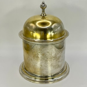 May include: A silver and gold metal container with a domed lid and a finial on top. The container has a smooth, polished surface.
