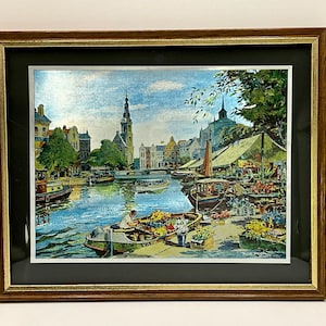 May include: A framed silver foil print of a canal scene in a European city. The print depicts a canal with boats, buildings, and a market with flowers and produce. The frame is brown wood with a gold trim.