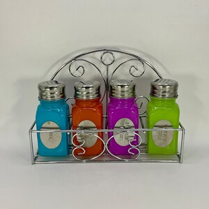 Op de afbeelding: Vier kleurrijke glazen kruidenpotten met zilveren deksels en etiketten, die op een zilveren metalen rek met een decoratief scrollontwerp staan.