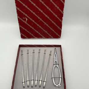 May include: A vintage set of silver cocktail forks and a silver tool, presented in a red box with a gold chain pattern. The forks are slender with a flat, rounded end. The tool has a long, pointed nose.