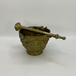 Puede incluir: Un mortero y un pilón de latón con un diseño ornamentado. El mortero es redondo y tiene una abertura ancha. El pilón es largo y delgado con un extremo redondeado.