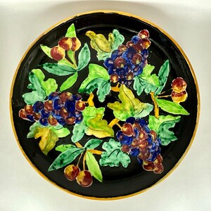 Könnte beinhalten: Ein schwarzer Keramikteller mit goldfarbenem Rand zeigt ein handgemaltes Design aus Trauben und Blättern. Die Trauben sind in Schattierungen von Lila, Blau und Grün bemalt, und die Blätter sind in Schattierungen von Grün und Gelb bemalt.