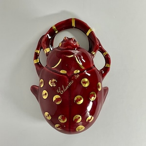 May include: A red ceramic beetle figurine with gold polka dots and a gold rim around the top. The beetle has a raised head and a small opening on the top. The figurine is signed "Clement Des" on the side.