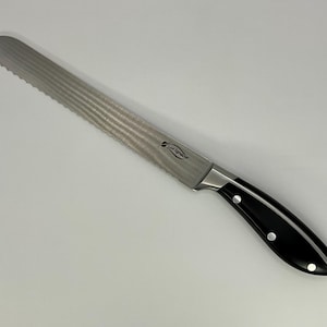 Puede incluir: Cuchillo de pan dentado con una hoja de acero inoxidable y un mango negro. El cuchillo tiene una punta curva y una virola plateada. El mango negro tiene tres remaches plateados. Diseñado para cortar pan.
