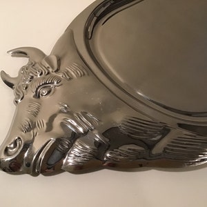 May include: A silver serving tray featuring a bull's head design. The tray is oval shaped and has a raised rim.