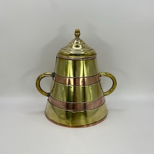 May include: A vintage brass container with a lid and two handles. The container has a gold finish with copper-colored bands around the body. The lid is also gold and has a decorative finial on top. The container is likely used for storage or display.