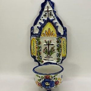 Op de afbeelding: Een wit keramisch wandgemonteerd wijwaterfonteintje met een blauw en geel geschilderd ontwerp. Het fonteintje heeft een kruis en bloemmotieven.