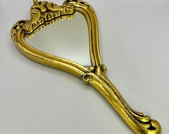 May include: A gold-colored hand mirror with an ornate, heart-shaped frame. The mirror has a long, curved handle with intricate details.
