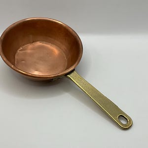 Peut inclure: Une petite casserole en cuivre avec une poignée en laiton. La casserole a une forme de bol arrondie et une longue poignée plate avec un trou à l'extrémité pour l'accrocher. Le cuivre a une surface brillante et réfléchissante.